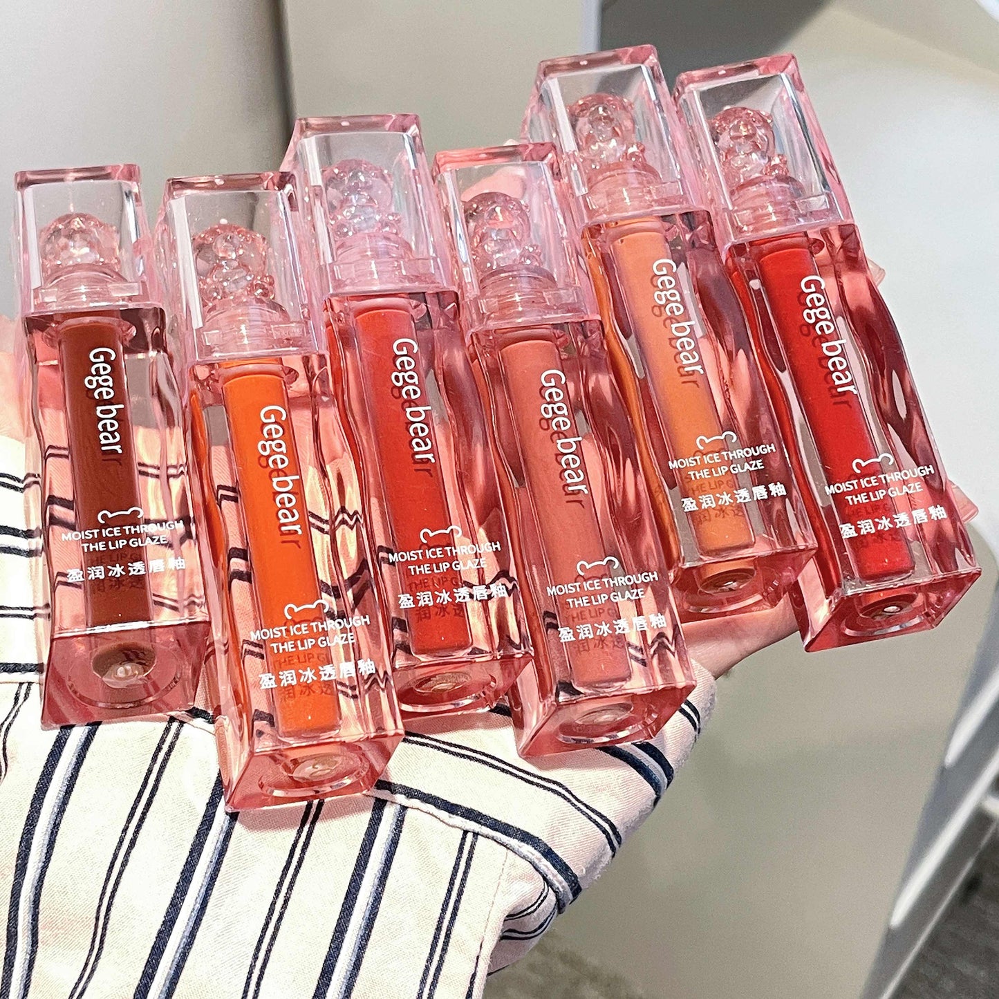 Multiple lip glosses with 'Gege bear' branding held by a person wearing a striped shirt.