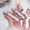 Hand holding a clear perfume bottle with silver cap, surrounded by lip products on a light background.