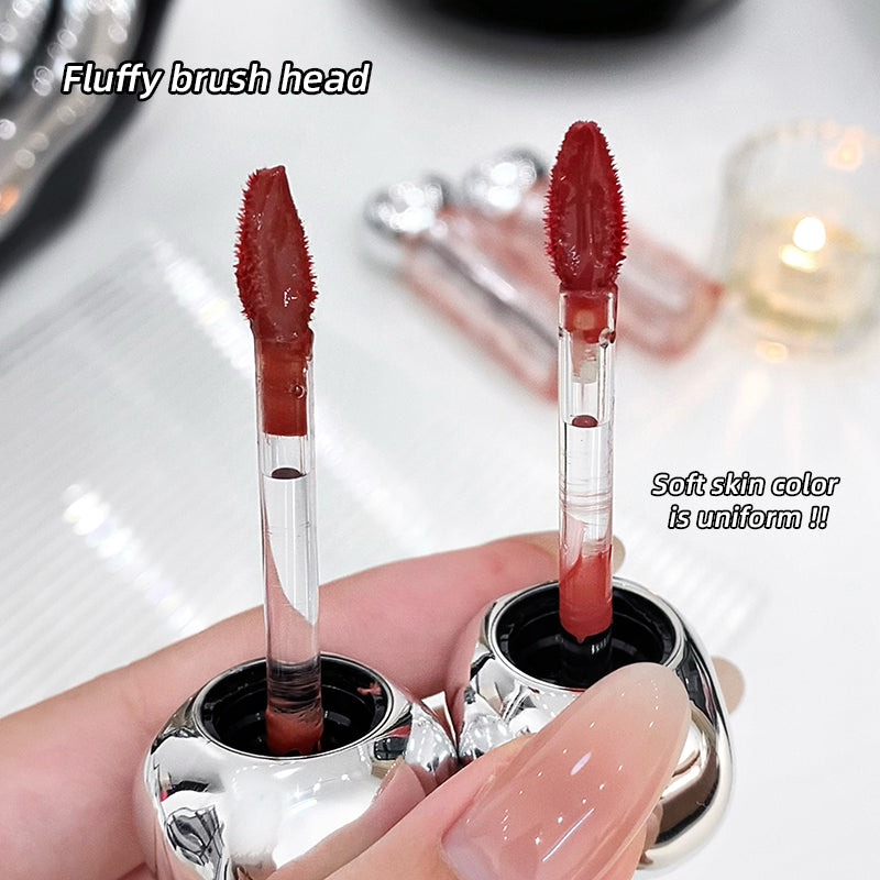 Two lip glosses with fluffy brush heads held by a hand against a blurred background.